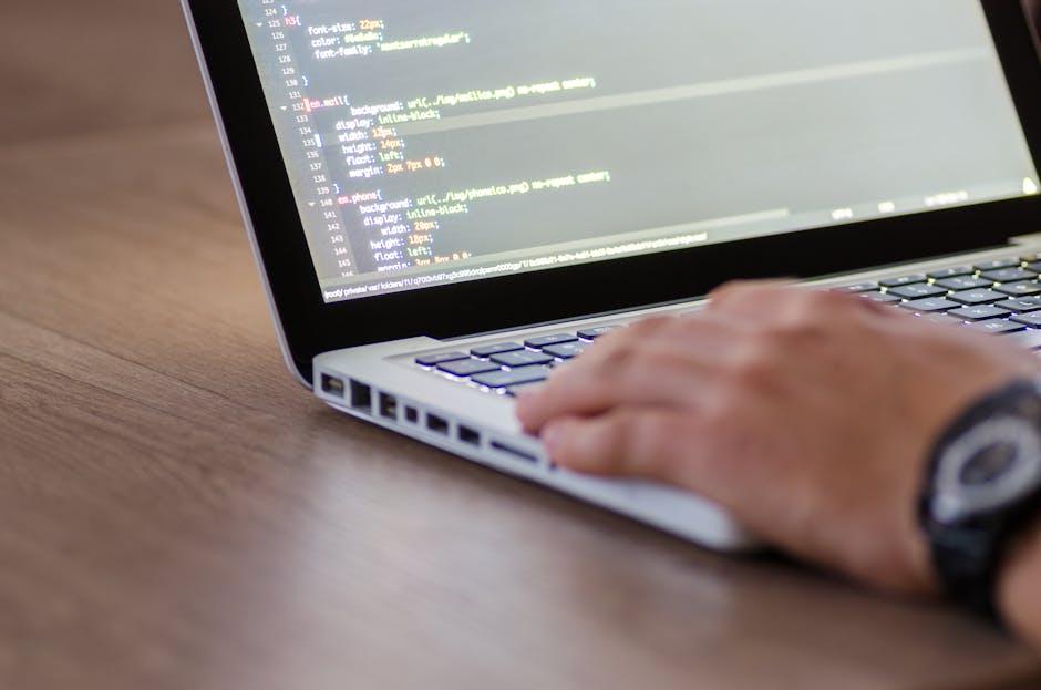 A close-up view of a person's hand operating a silver laptop on a wooden desk, with the screen displaying lines of colorful programming code. The person's wristwatch is partially visible on their left
